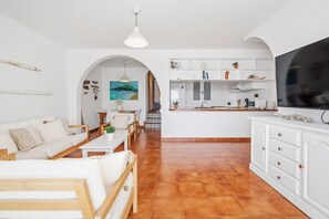Un salon comprenant un canapé, une table basse et un téléviseur, attenant à une cuisine avec un coin repas et donnant sur la mer.