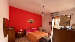 A bedroom with a red wall, a bed with an orange bedspread, a wooden nightstand with a lamp, a chair, a desk with a computer, and a fan on the wall. - Emily Home (Giardini Naxos)