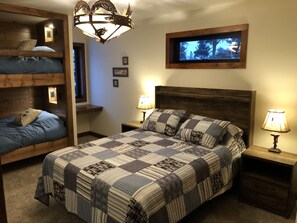 Un dormitorio con una cama grande, dos mesitas de noche, un cabecero de madera, un televisor montado en la pared y una cama litera a la izquierda.