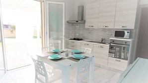 Une cuisine moderne avec des meubles blancs, un four à micro-ondes et une cuisinière à gaz. On y trouve une table à manger avec des chaises et une porte vitrée donnant sur un espace extérieur.