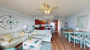 A living room with a sofa, coffee table, and a dining area with chairs. There is a kitchen in the background with wooden cabinets and a microwave.