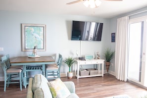 A living room with a wooden dining table, teal chairs, a sofa, a flat-screen TV mounted on the wall, and a sliding glass door.