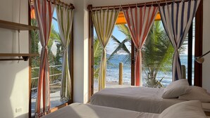 A bedroom with two beds, large windows with colorful curtains, a view of the beach and palm trees, and a wooden headboard. - Casa Naranja: Ocean View (Puerto Villamil)