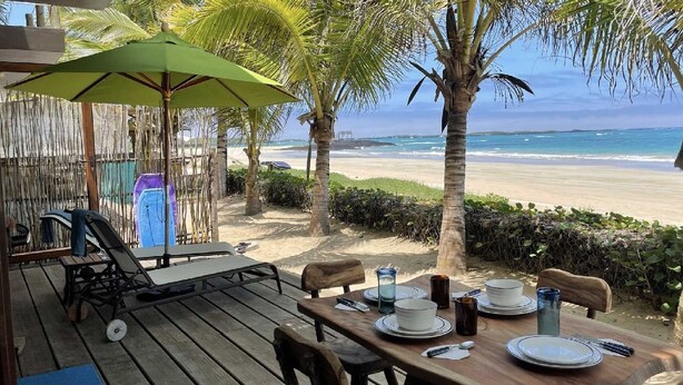 A wooden deck with a table set for dining, overlooking a beach with palm trees and a clear view of the ocean. - Casa Naranja: Ocean View (Puerto Villamil)