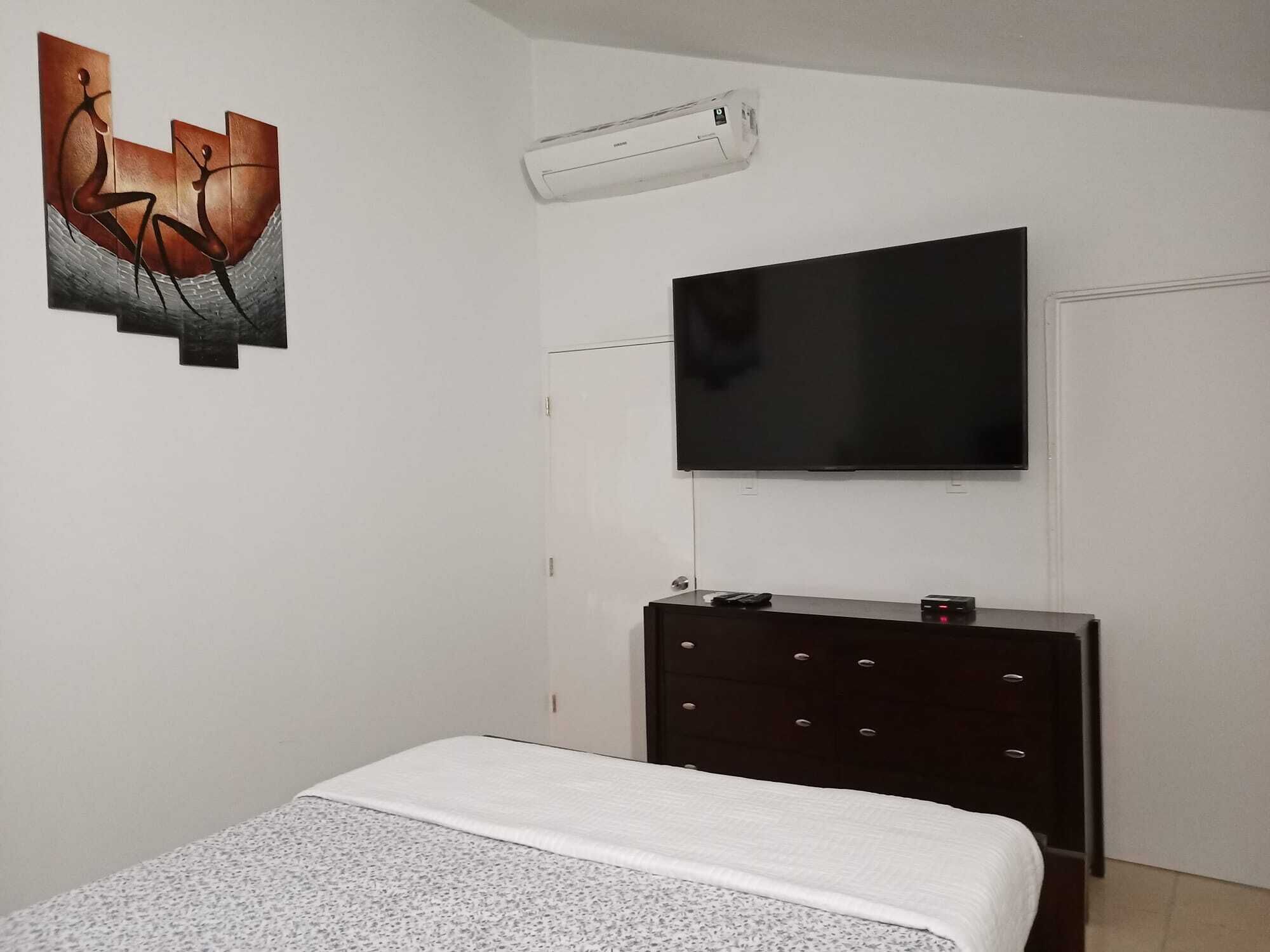 A bedroom with a bed, a dresser, a TV mounted on the wall, and an air conditioning unit.