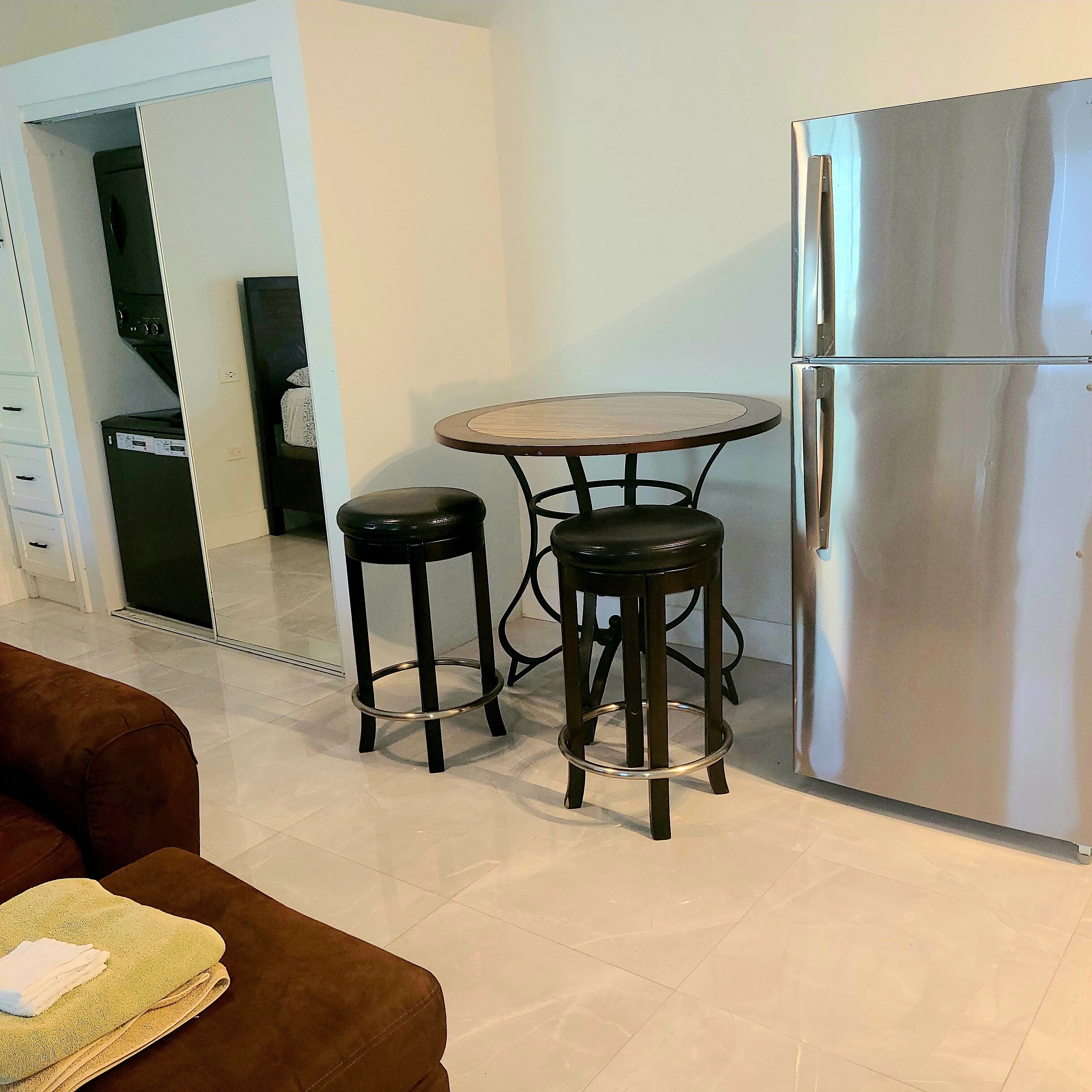A compact kitchen with a stainless steel refrigerator, a small round table with two stools, and a brown sofa with a towel.