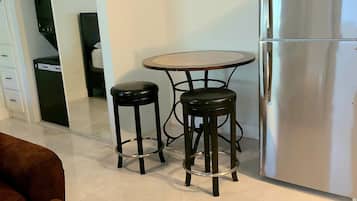 A compact kitchen with a stainless steel refrigerator, a small round table with two stools, and a brown sofa with a towel.