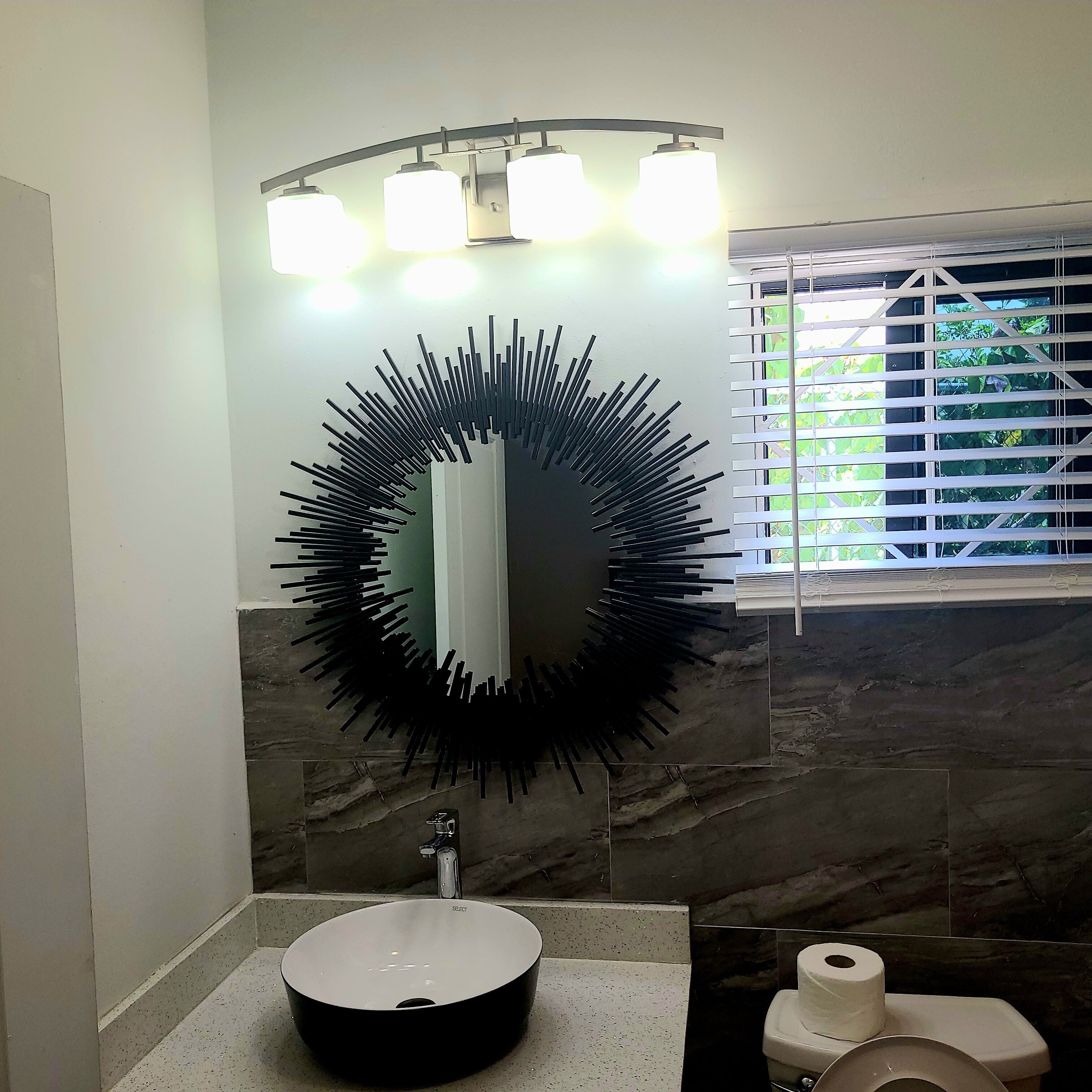 Een badkamer met een ronde spiegel, een wastafel en een toilet. Er is een raam met zonwering en een lamp boven de spiegel.