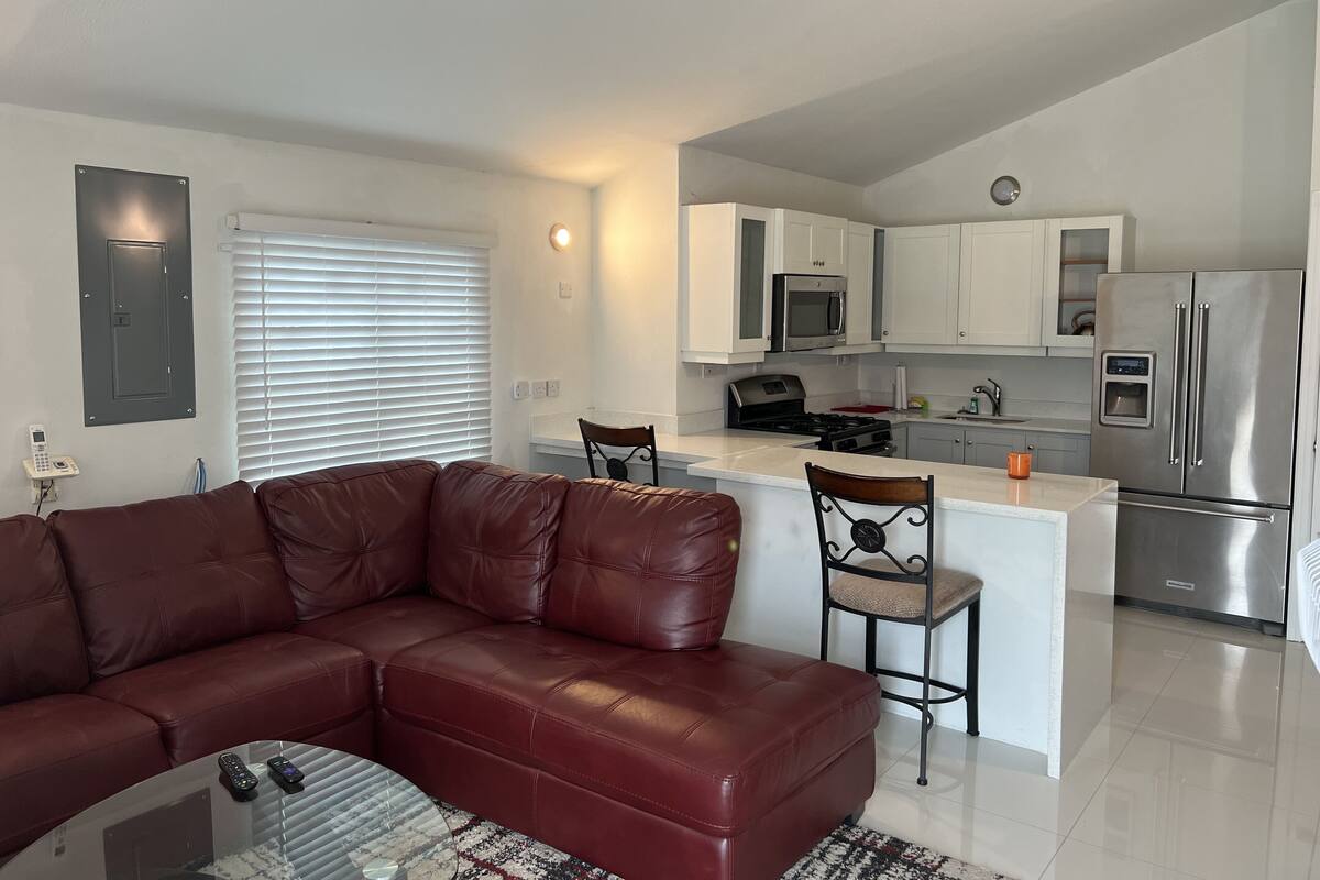 A modern living room with a brown leather sofa, a glass coffee table, and a kitchen area featuring white cabinets, stainless steel appliances, and a small island with bar stools.