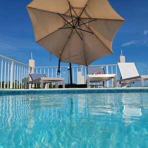 A swimming pool with clear blue water, white lounge chairs, and a large beige umbrella providing shade. - Villa Jamaica (Falmouth)