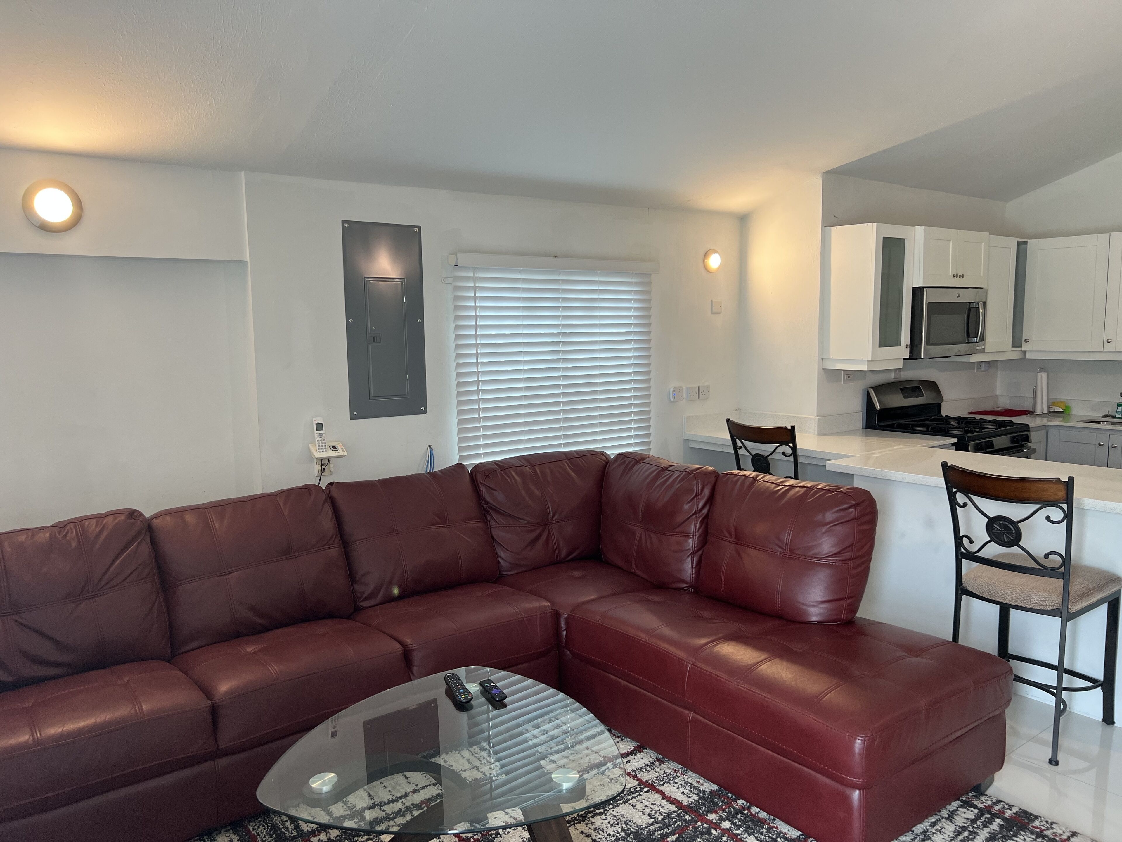 A living room with a brown leather sofa, a glass coffee table, and a kitchen area with white cabinets and appliances.