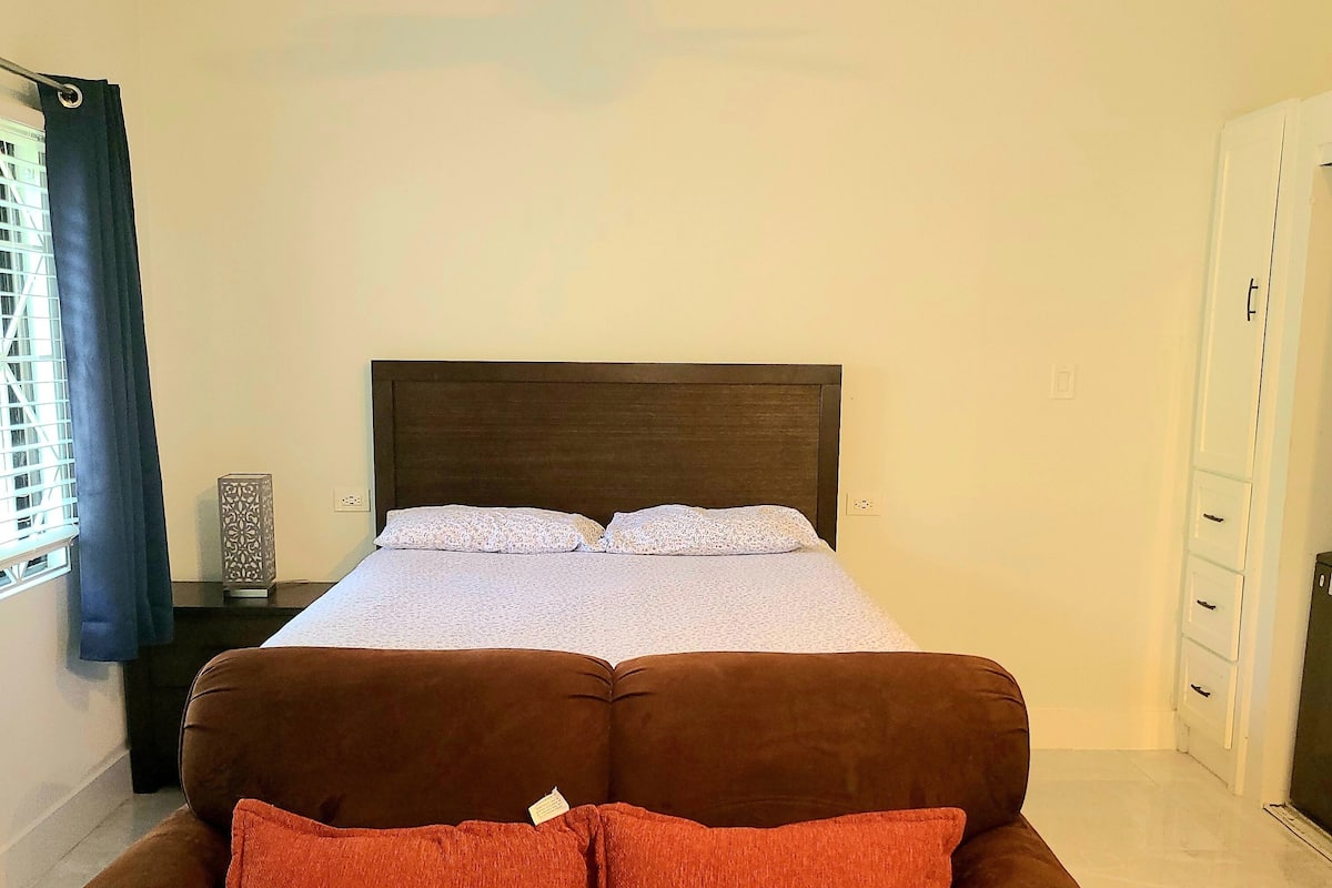 A bedroom with a bed, a nightstand, a dresser, and a brown sofa with red cushions.