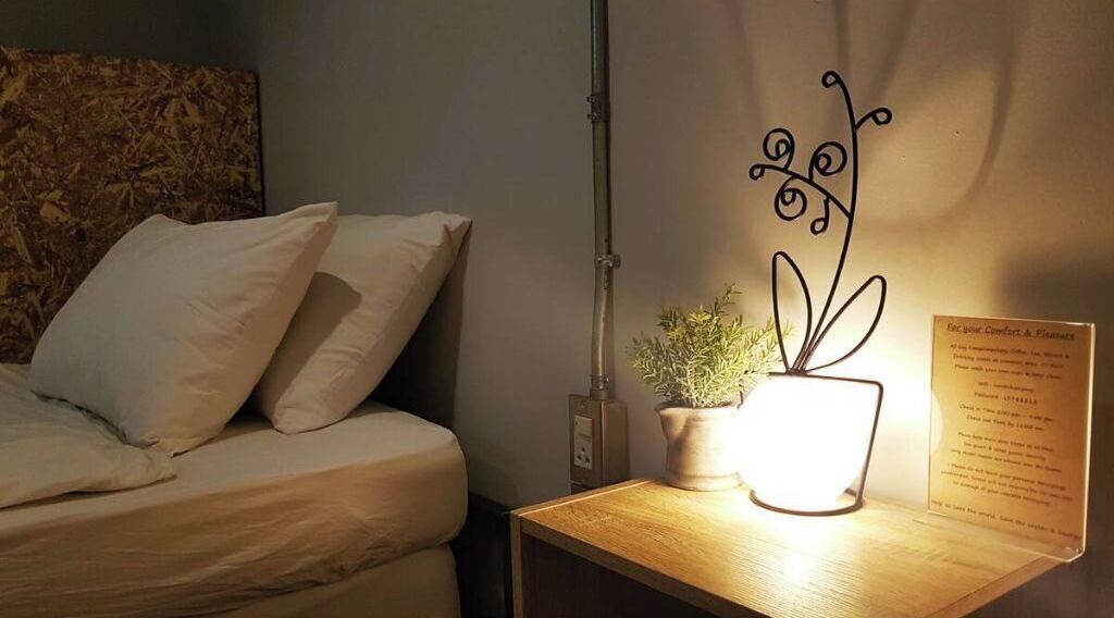 Een net opgemaakt bed met witte lakens, een klein nachtkastje met een potplant en een lamp, en een bord op de muur.