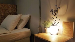 Ein ordentlich gemachtes Bett mit weißer Bettwäsche, ein kleiner Nachttisch mit Topfpflanze und Lampe sowie ein an der Wand befestigtes Schild.