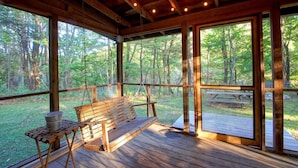 A wooden deck with a bench and a table, large windows offering a view of a forest.