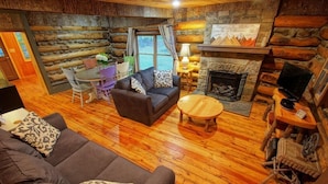 A log cabin interior with a wooden floor, a sofa, a table, a fireplace, and a stone wall.