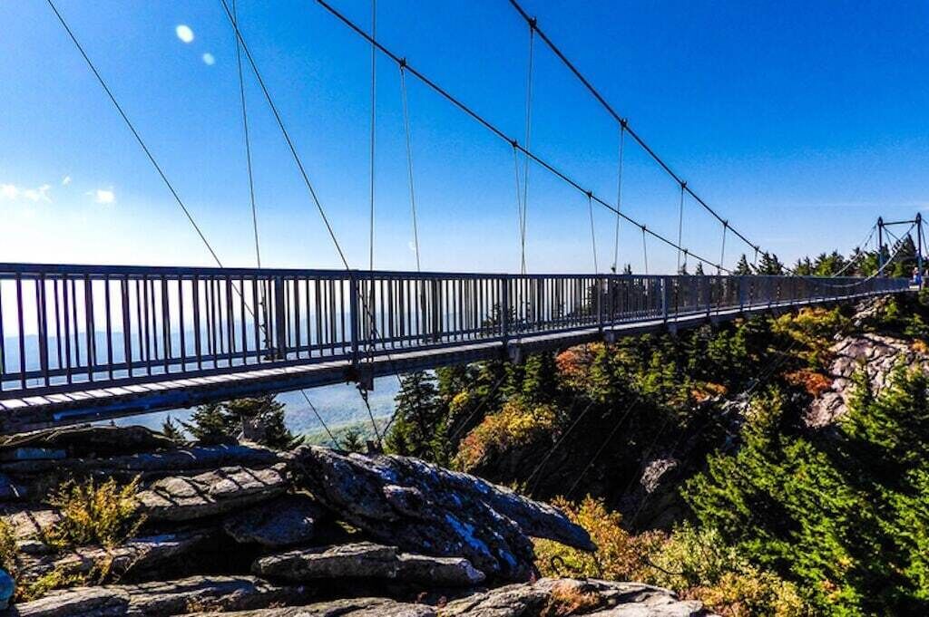 A suspension bridge with metal railings and cables, surrounded by rocky terrain and trees.