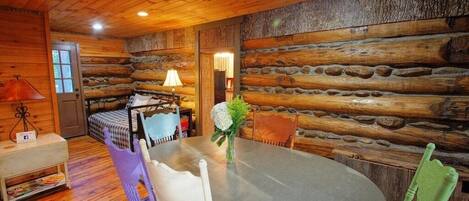 A log cabin interior with a round dining table, two chairs, a small sofa, a lamp, and a door.