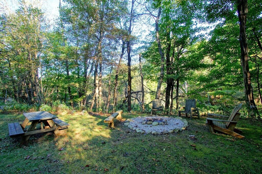 A forested campsite with a picnic table, benches, and a fire pit.