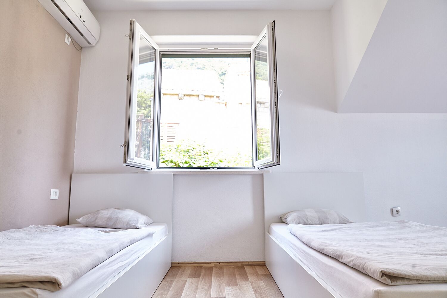 A small, bright room with two beds, a window, and an air conditioning unit.