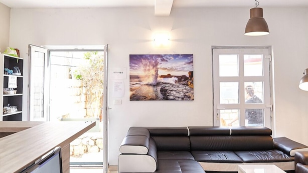 A modern living room with a black leather sofa, a wooden coffee table, and a laptop on a desk. There is a bookshelf with shoes and a painting on the wall.