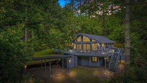 A modern cabin with a deck and a covered patio, surrounded by dense forest.
