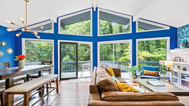 A spacious living room with a high ceiling, large windows, and a wooden dining area.