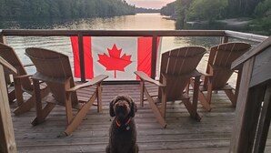 A wooden deck with three wooden chairs facing a river, a Canadian flag, and a dog in the foreground. - Muskoka Waterfront Cottage on 54 acres, 10 minutes from Gravenhurst (Gravenhurst)