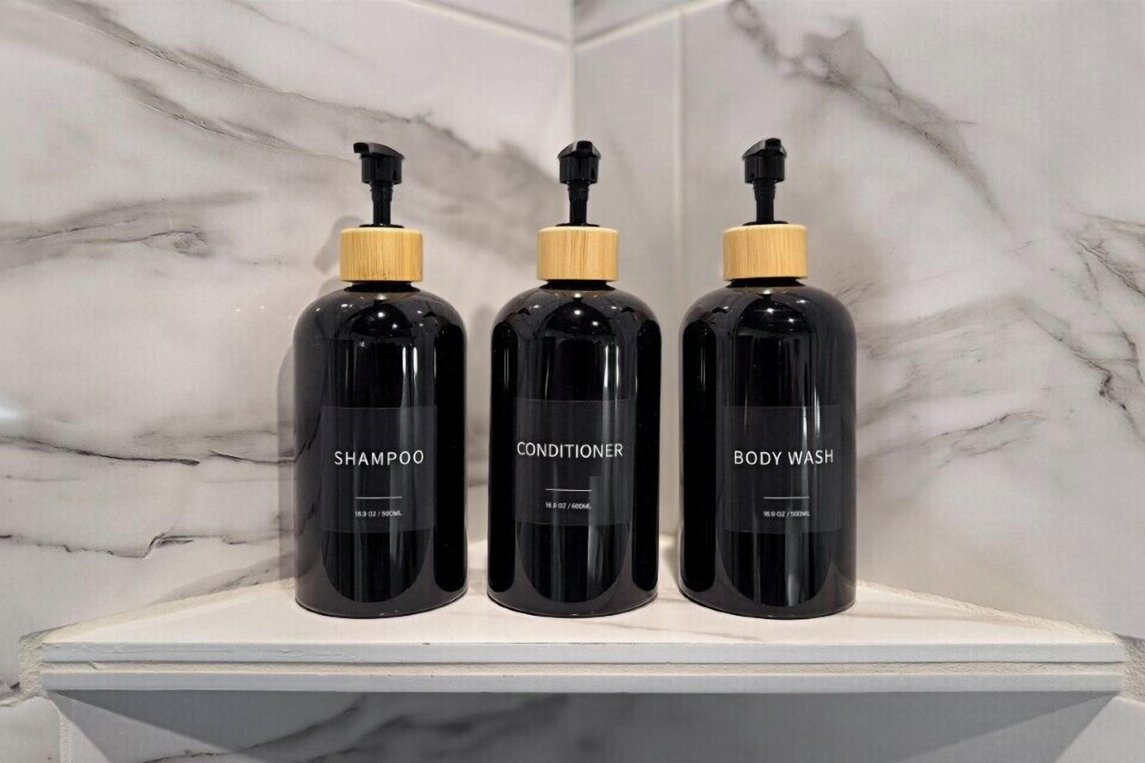 Three black bottles with wooden caps labeled 'SHAMPOO,' 'CONDITIONER,' and 'BODY WASH' on a white shelf against a marble wall.