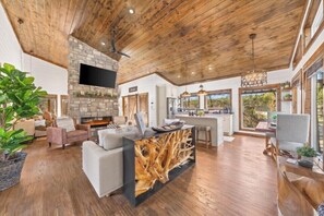 A spacious living area with a stone fireplace, wooden ceiling, and large windows offering views of the outdoors. - Private Pool · Sauna · Hot Tub · Just for Two (Broken Bow)