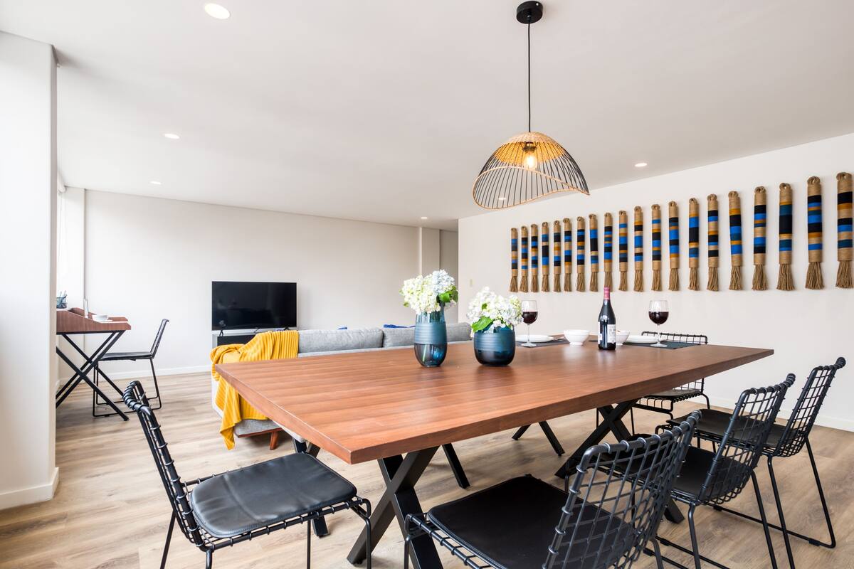 A modern dining area with a wooden table, black chairs, a yellow tablecloth, and a TV in the background.