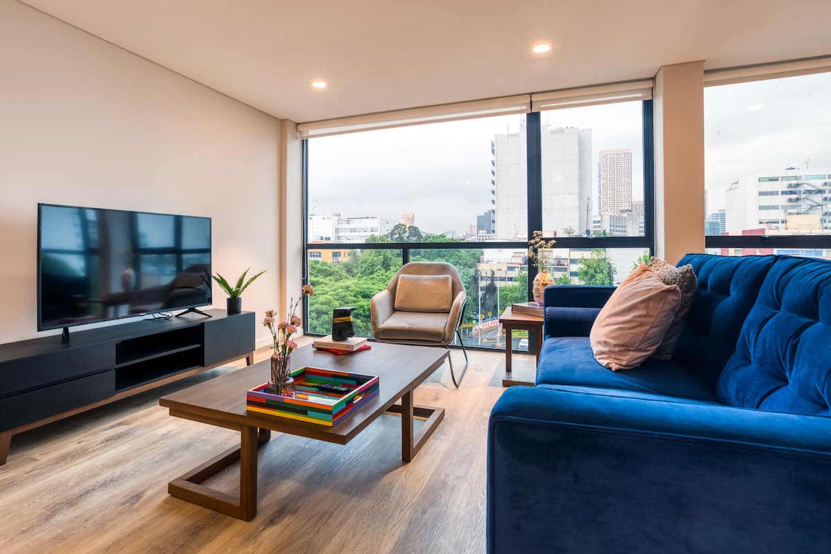 A modern living room with a large TV, a blue sofa, a coffee table, and a city view through large windows.