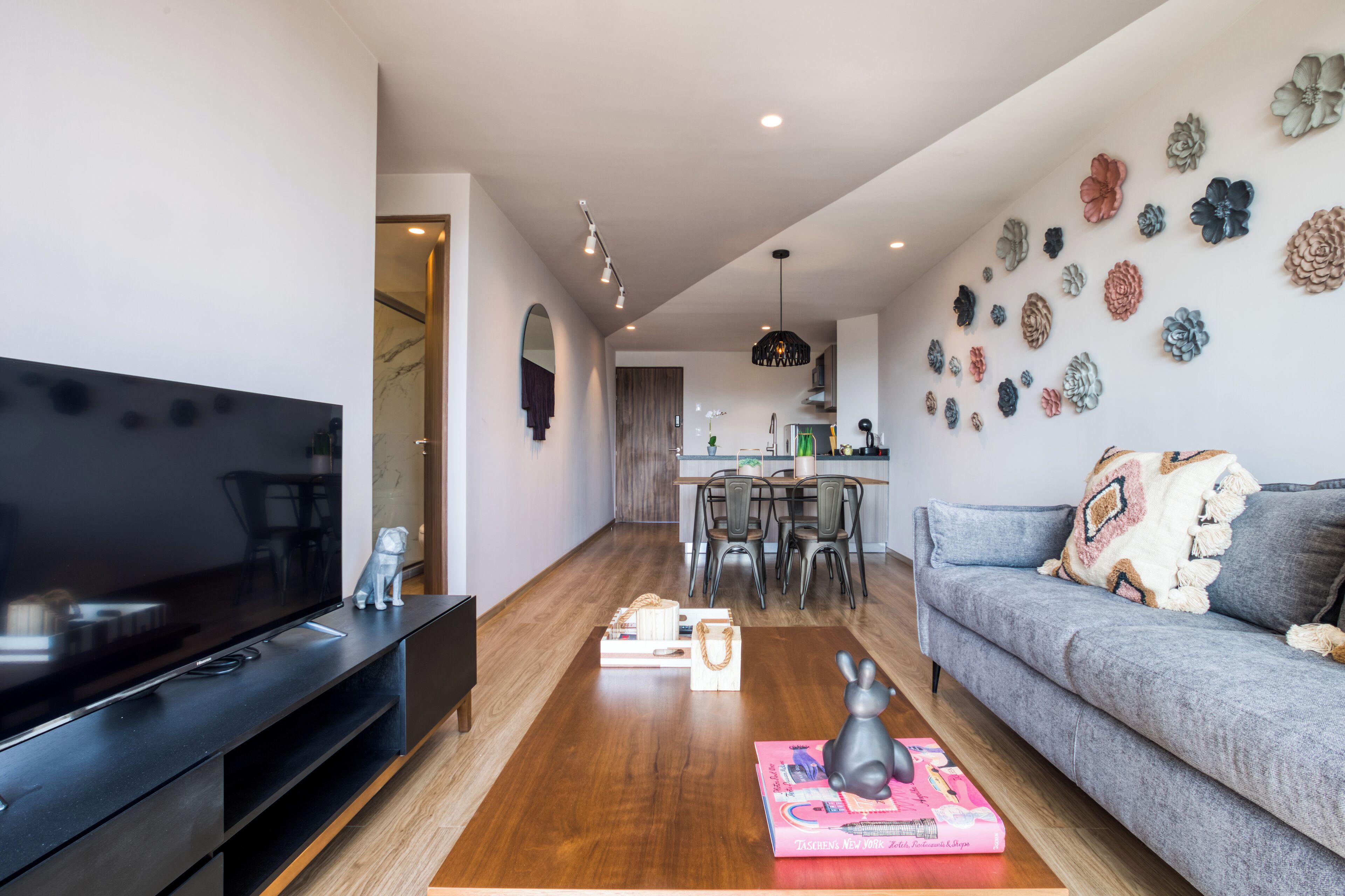 A modern living room with a flat-screen TV, a grey sofa, a wooden coffee table, and a dining area with chairs and a kitchen in the background.