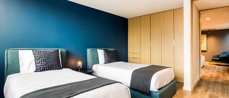 Moderni hotellihuone, jossa on kaksi sänkyä, sininen tehosteseinä, puulattia ja näkymä käytävään.