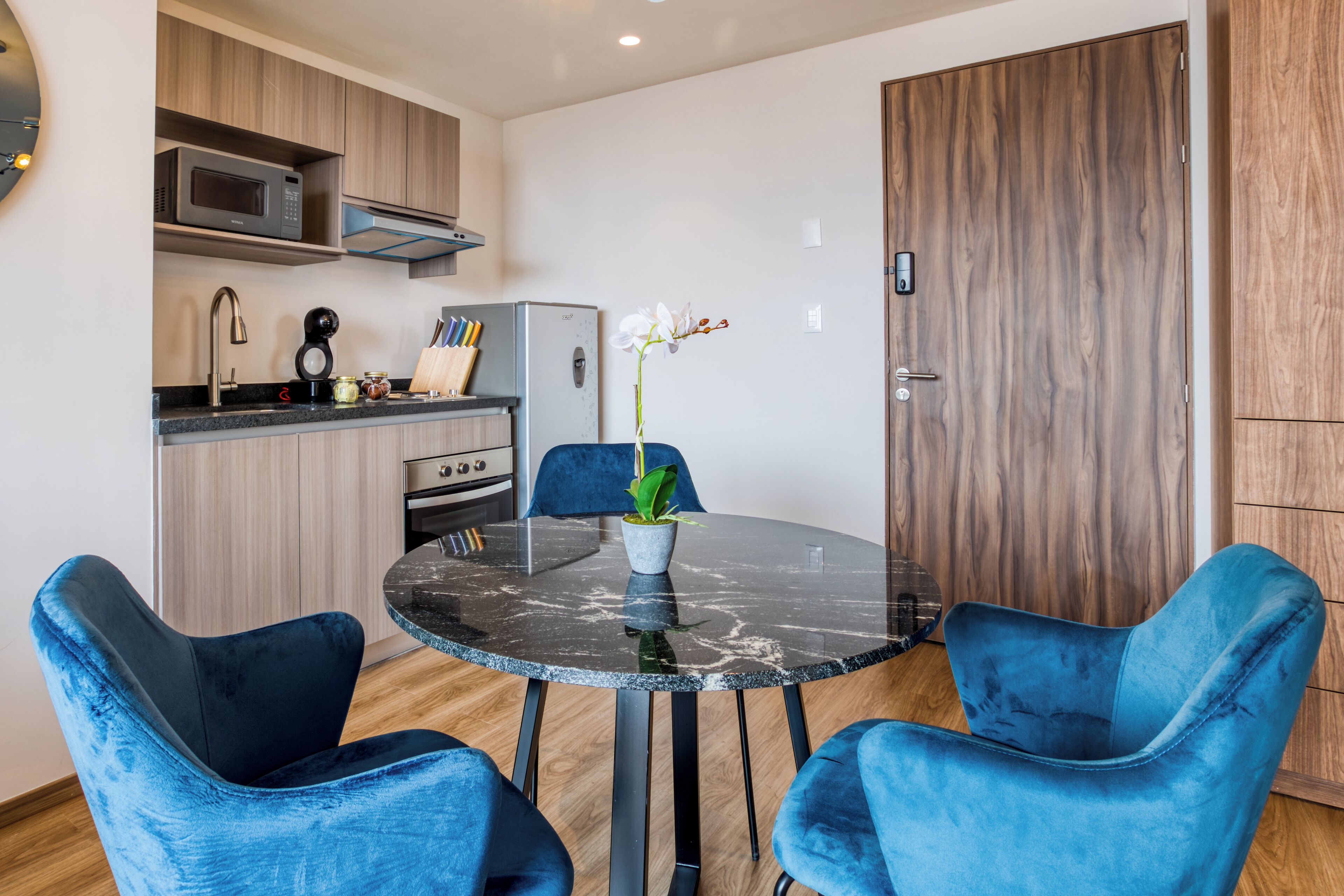 A compact kitchen with modern appliances, a dining table with two blue chairs, and a wooden door.