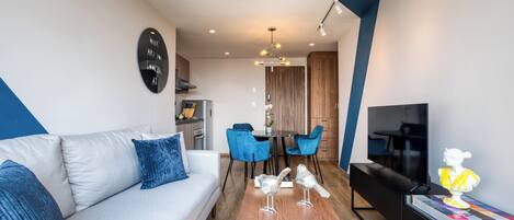 A modern living room with a sofa, a coffee table, and a TV. There is a dining area with chairs and a table.