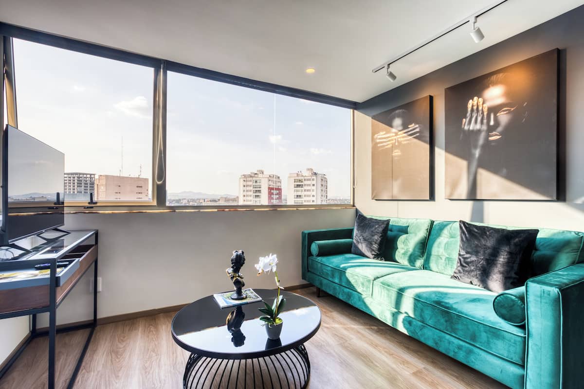 A modern living room with a green sofa, a coffee table, and a large window offering a city view.