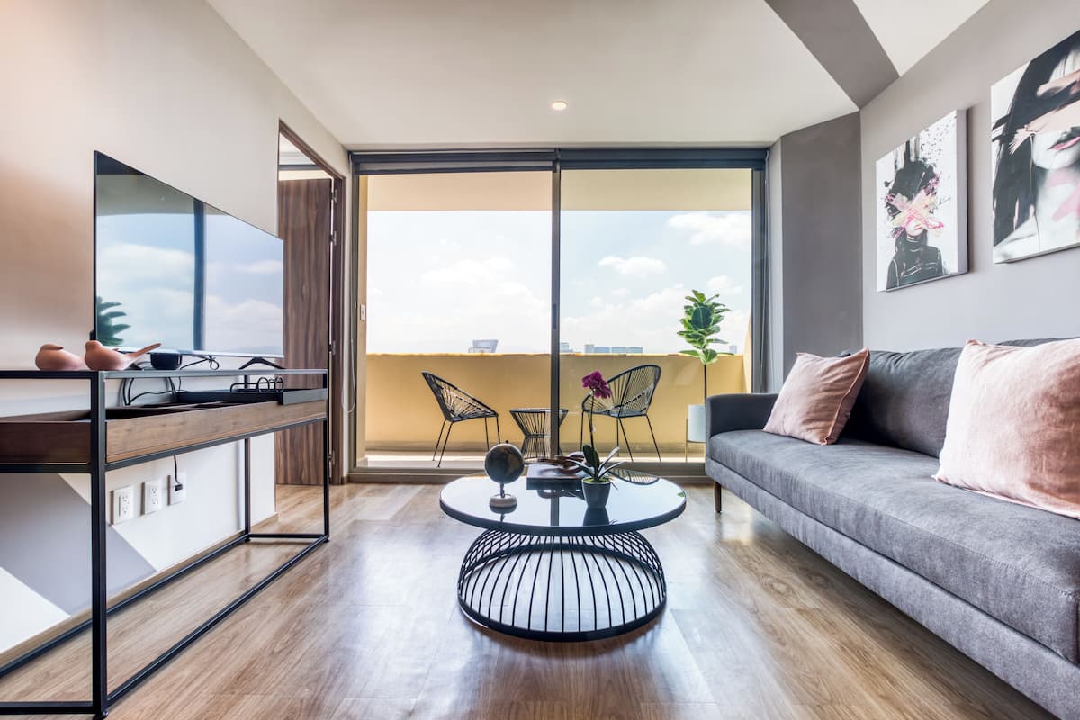 A modern living room with a grey sofa, a round coffee table, and a TV on a wooden cabinet. There are two chairs and a plant on the table.