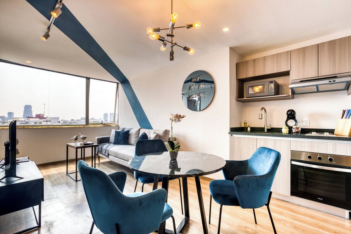 A modern kitchen with a microwave, stove, and sink. There is a dining area with a round table and blue chairs. A large window offers a view of the city skyline.