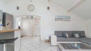 Een moderne woonruimte met een keuken, een grijze bank met kussens, een salontafel en een spiltrap die naar een hoger niveau leidt.