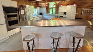 A modern kitchen with stainless steel appliances, a large island with bar stools, and wooden paneling on the walls. - North House - over 100 acres of Adirondack privacy! (Jay)