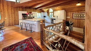 A spacious kitchen with wooden beams, a central island with stools, and a rustic staircase. - North House - over 100 acres of Adirondack privacy! (Jay)
