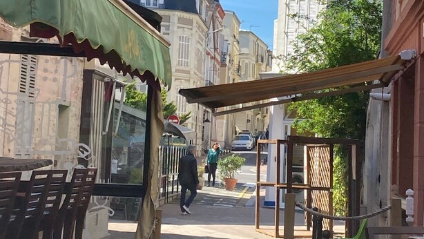 A narrow street with buildings on either side, a covered seating area with chairs, and a person walking in the distance.