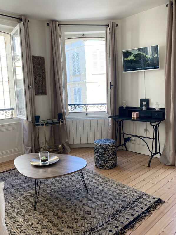 A room with a round table, a leopard print ottoman, a radiator, a TV on the wall, and a window with curtains.