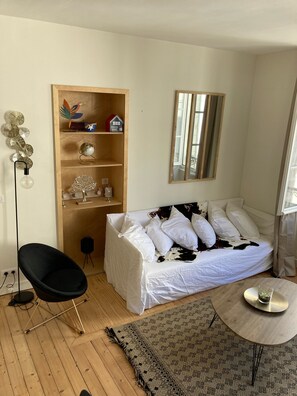 A bedroom with a bed, a chair, a small table, a mirror, and a wooden shelf with decorative items.