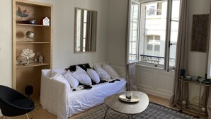 A cozy room with a white sofa, a small round table, a wooden shelf with decorative items, and a large window with curtains.