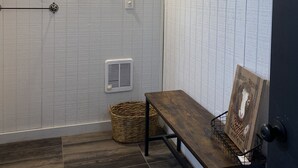 Una habitación con techo de madera, pared blanca con textura, un cuadro enmarcado, un banco, una cesta de mimbre y un estante pequeño.