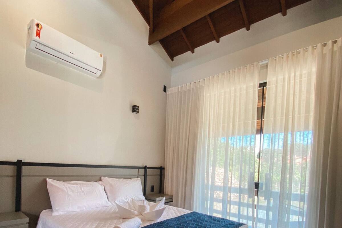 Habitación de hotel con cama, cortinas blancas, unidad de aire acondicionado y vistas por la ventana.