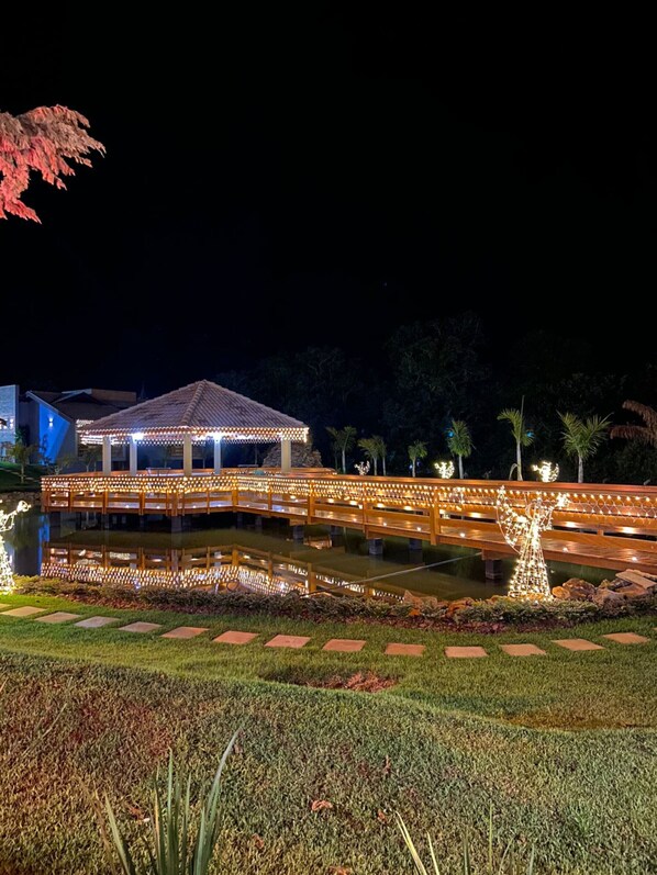 A nighttime view of a well-lit gazebo over a reflective pond, surrounded by landscaped gardens and palm trees. - Pousada Saint Diniz (Monte Sião)