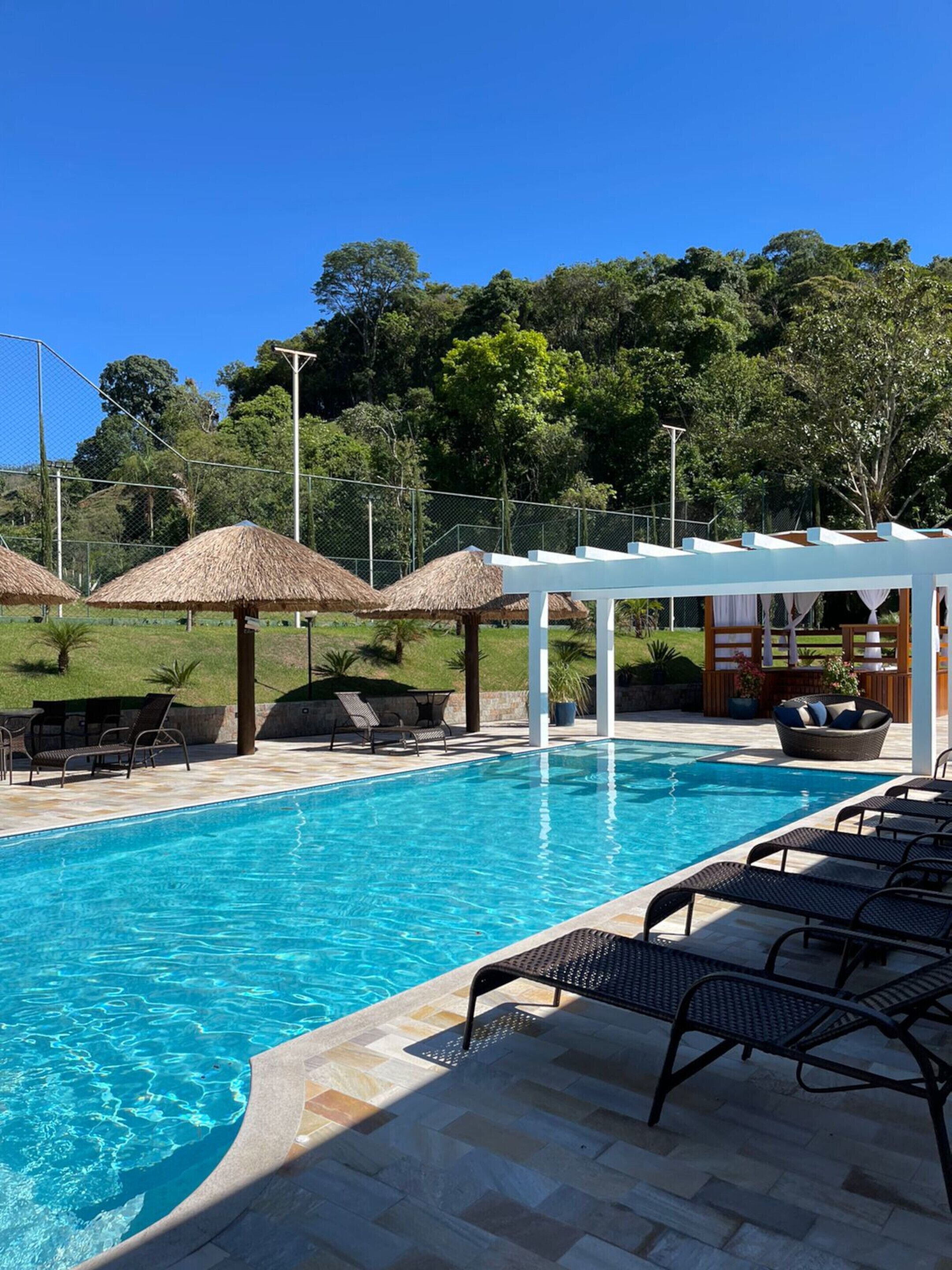 A swimming pool with lounge chairs and thatched umbrellas, surrounded by a fence and greenery.